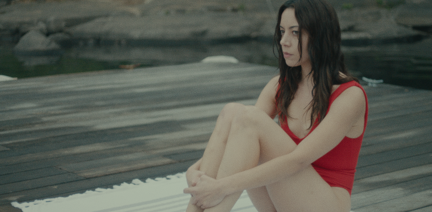 Aubrey Plaza wearing a red bathing suit sitting on a dock in a still from Black Bear