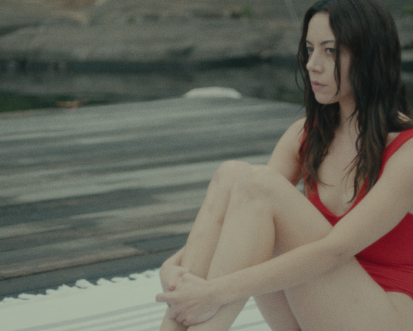 Aubrey Plaza wearing a red bathing suit sitting on a dock in a still from Black Bear
