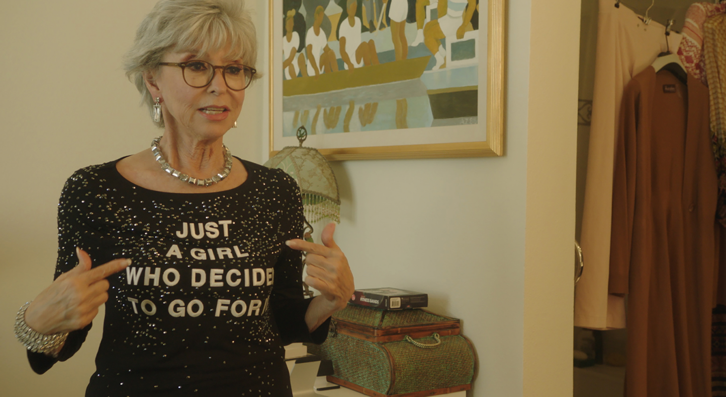 Rita Moreno pointing to her shirt, which say Just A Girl Who Decided to Go For It