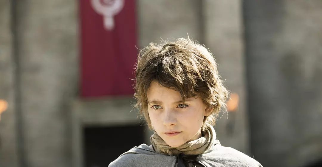 A young girl with a boy's haircut poses in front of a medieval castle