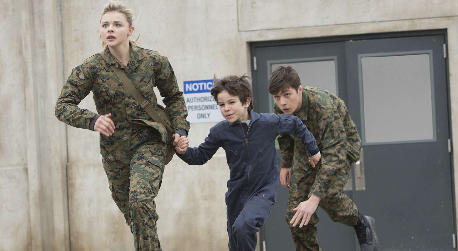 Cassie (Chloë Grace Moretz) and Ben (Nick Robinson), wearing army fatigues, run with pre-teen Sam (Zachary Arthur)
