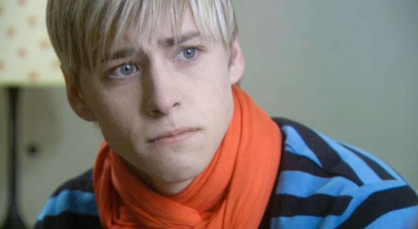 Mitch Hewer as Maxxie, wearing a blue and black striped shirt and red scarf