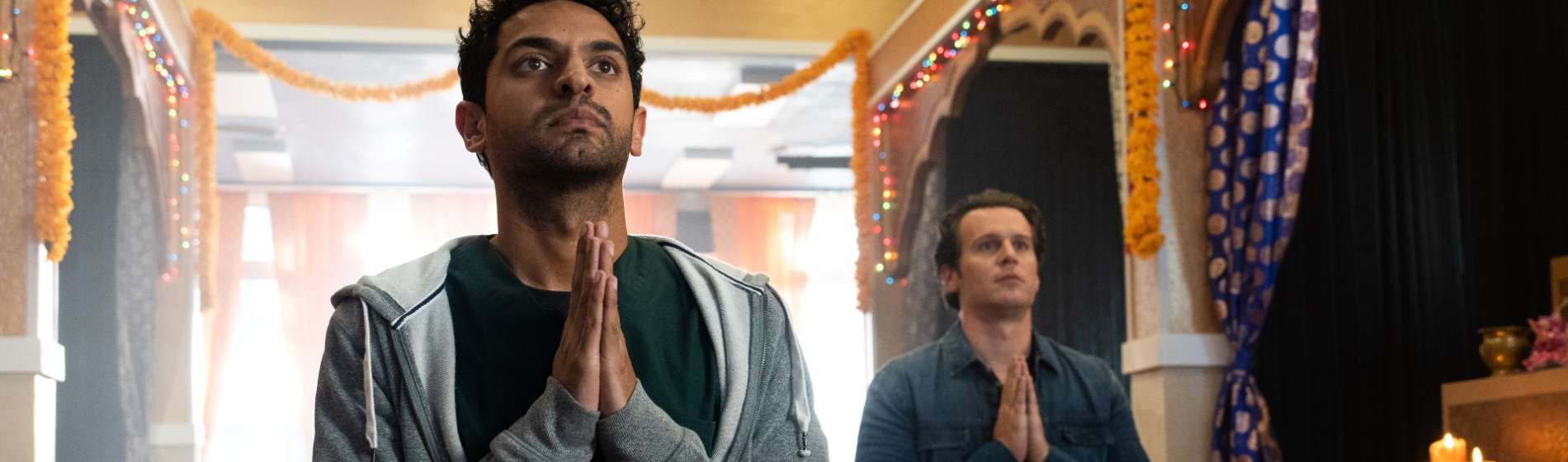 An Indian man in a sweat hoodie and a white man in a blue jacket kneel and pray on a red carpet in a temple.