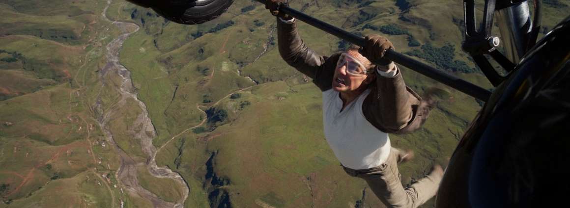 Tom Cruise dangles from an airplane. He is hanging high above a field, and wearing a tan suit, white shirt, and goggles. There is a lot of wind in his hair.