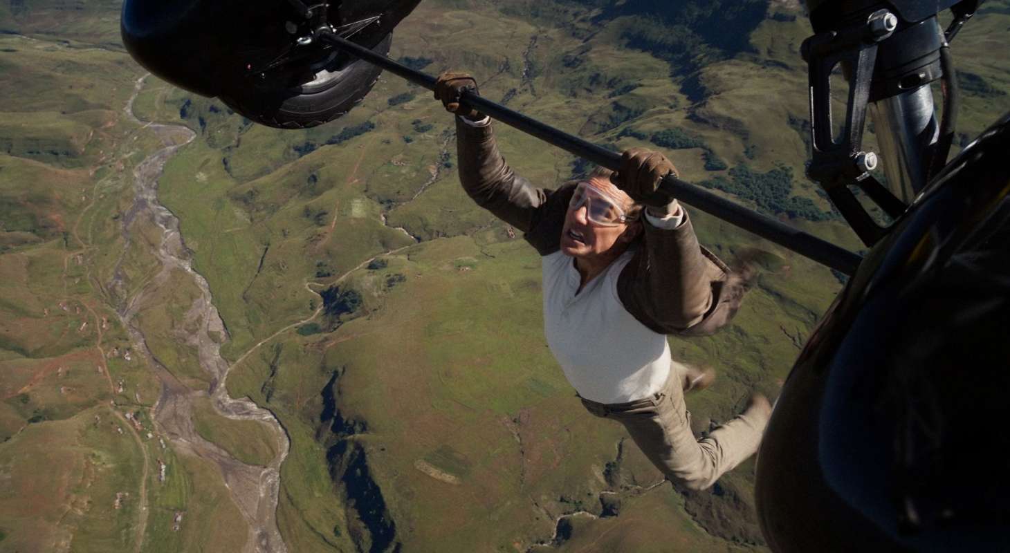 Tom Cruise dangles from an airplane. He is hanging high above a field, and wearing a tan suit, white shirt, and goggles. There is a lot of wind in his hair.