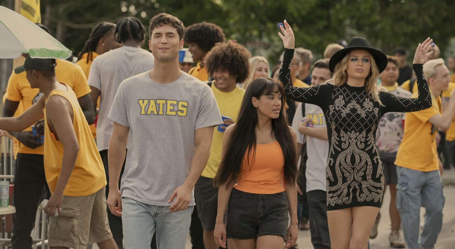 A tall young man in a grey t-shirt that says Yates in yellow letters walks in a crowd with a shorter young woman wearing an orange tank top and black skirt. A girl beside them wears a quasi-witch outfit and has her hands in the air.
