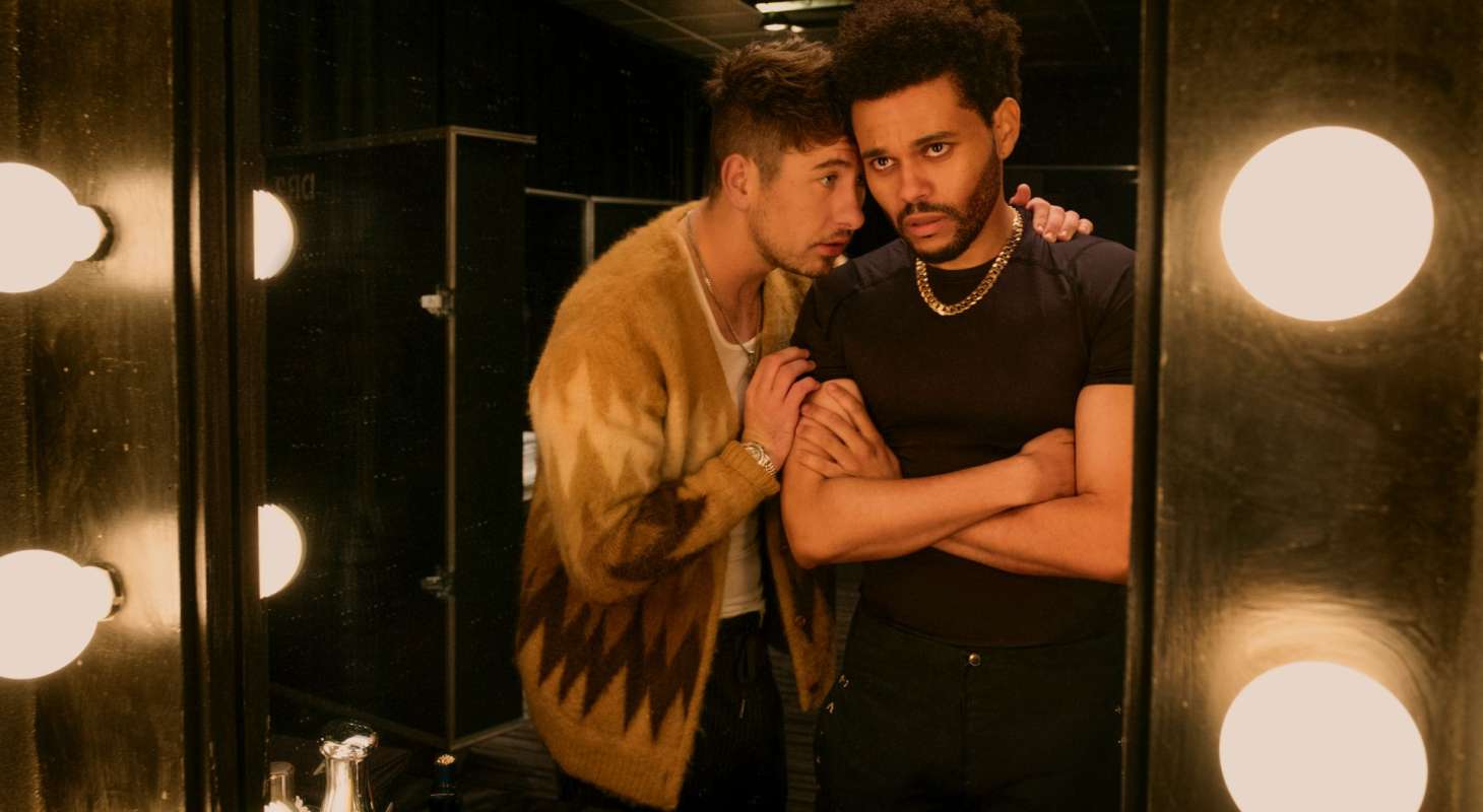 Actors Barry Keoghan and Abel Tesfaye (The Weeknd) stand looking into a mirror. Keoghan is wearing a gold and brown sweater and Tesfaye is wearing a black t-shirt with a gold chain.