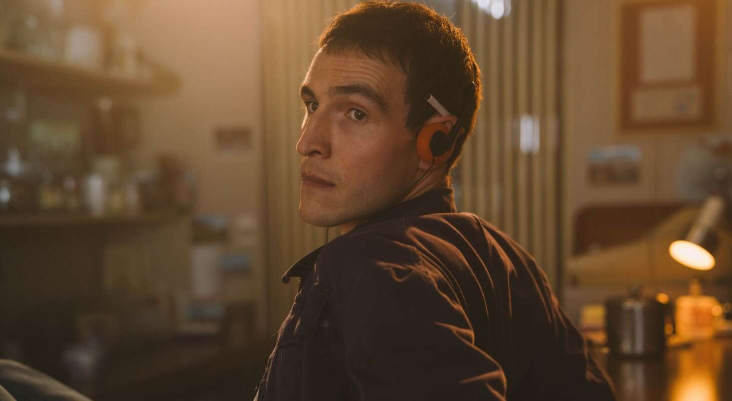 A young Irishman with brown hair and brown eyes is pictured from the waist up, leaning against a bar and looking towards screen left at someone outside the frame. He has orange headphones and a cigarette tucked under his ear.