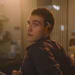 A young Irishman with brown hair and brown eyes is pictured from the waist up, leaning against a bar and looking towards screen left at someone outside the frame. He has orange headphones and a cigarette tucked under his ear.