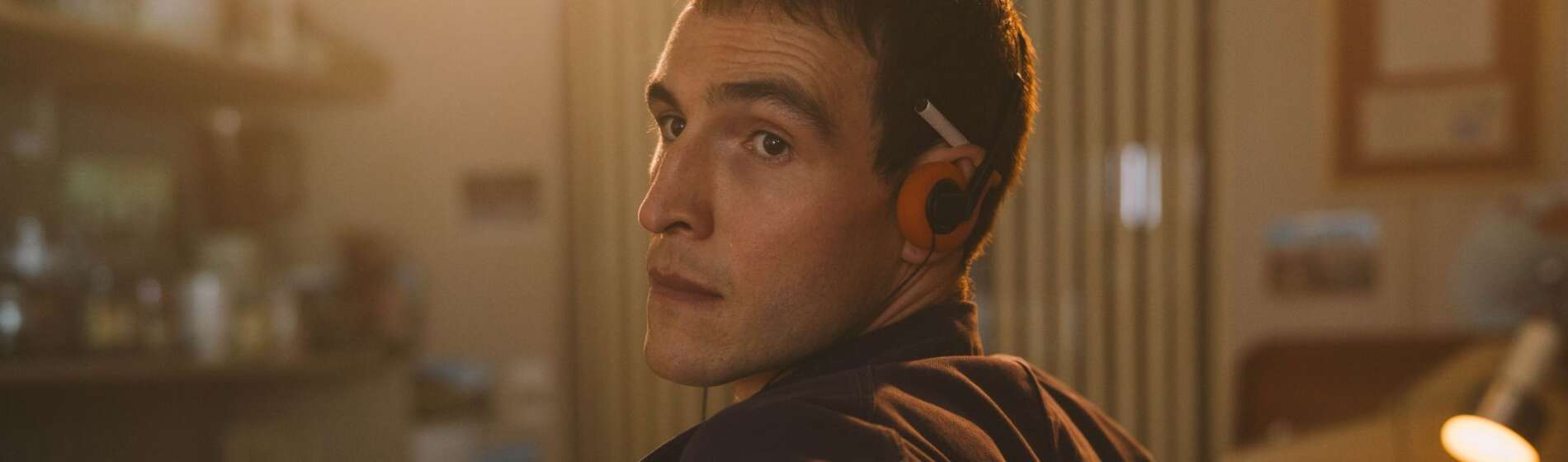 A young Irishman with brown hair and brown eyes is pictured from the waist up, leaning against a bar and looking towards screen left at someone outside the frame. He has orange headphones and a cigarette tucked under his ear.
