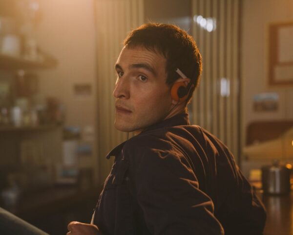 A young Irishman with brown hair and brown eyes is pictured from the waist up, leaning against a bar and looking towards screen left at someone outside the frame. He has orange headphones and a cigarette tucked under his ear.