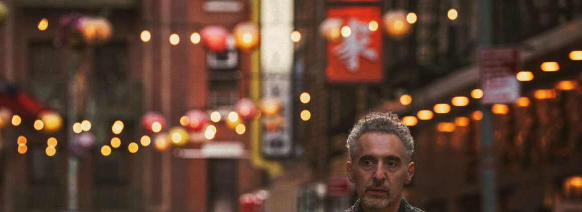 John Turturro stands in the foreground of a image of New York City's Chinatown neighbourhood, with street signs and lanterns behind him.
