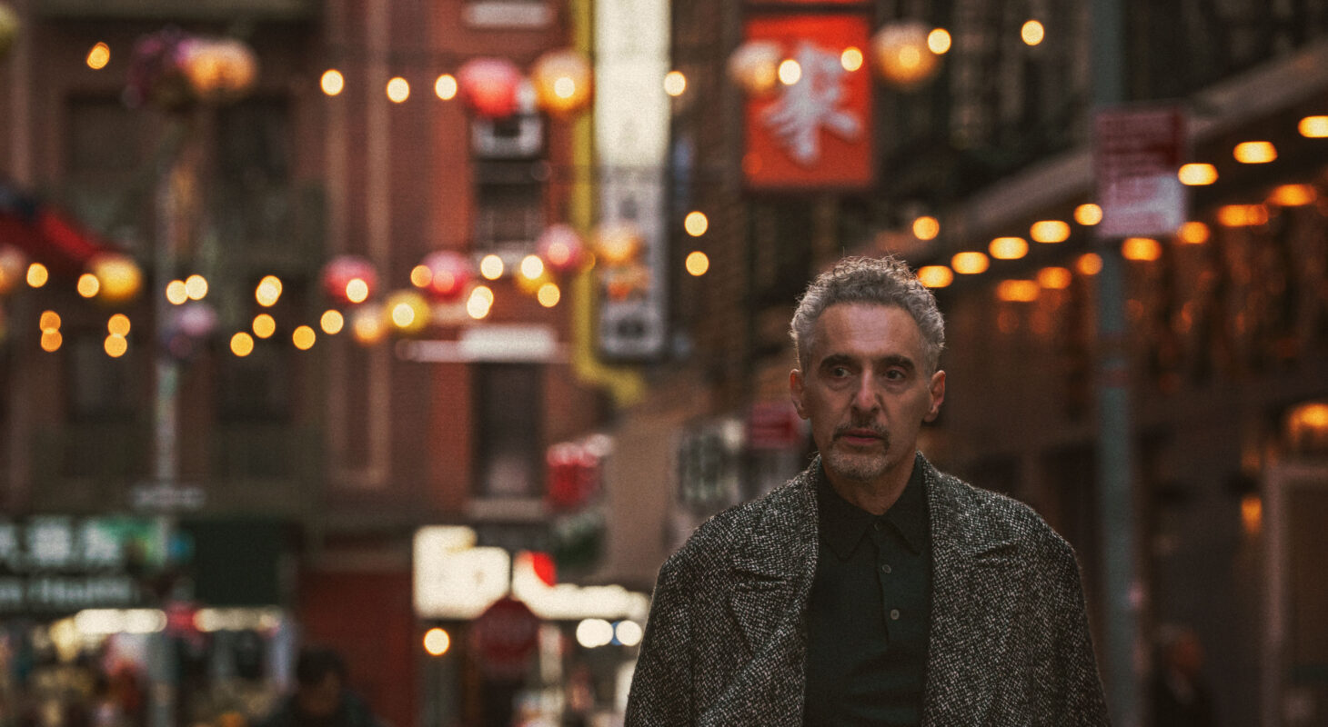 John Turturro stands in the foreground of a image of New York City's Chinatown neighbourhood, with street signs and lanterns behind him.