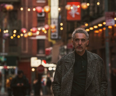 John Turturro stands in the foreground of a image of New York City's Chinatown neighbourhood, with street signs and lanterns behind him.