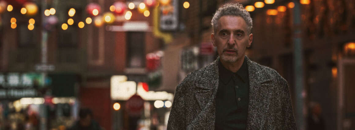 John Turturro stands in the foreground of a image of New York City's Chinatown neighbourhood, with street signs and lanterns behind him.
