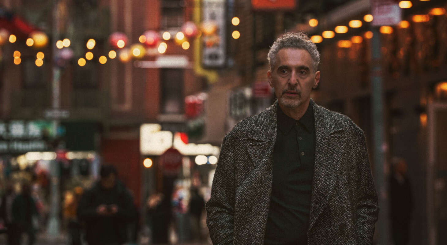 John Turturro stands in the foreground of a image of New York City's Chinatown neighbourhood, with street signs and lanterns behind him.