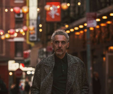John Turturro stands in the foreground of a image of New York City's Chinatown neighbourhood, with street signs and lanterns behind him.