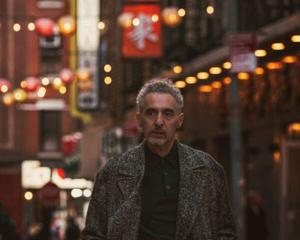 John Turturro stands in the foreground of a image of New York City's Chinatown neighbourhood, with street signs and lanterns behind him.