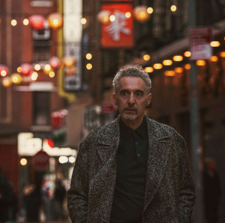 John Turturro stands in the foreground of a image of New York City's Chinatown neighbourhood, with street signs and lanterns behind him.
